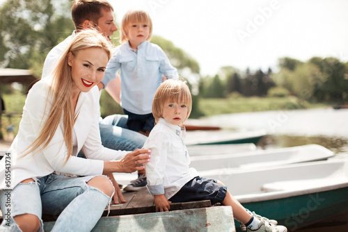 Family of four - parents and two twin boys sitting on pier near boats outdoors in park in sunny weather. Celebrating holidays and happy time together. Outdoor family activity, leisure time together