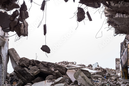 A hole in the body of a building with a pile of construction debris and concrete fragments hanging on the rebar against a uniform gray sky Fototapet