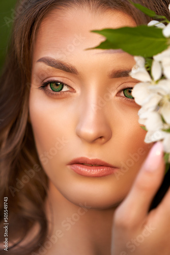 Portrait of a green-eyed girl with a branch of a flowering apple tree in her hand near her cheek. Close-up.