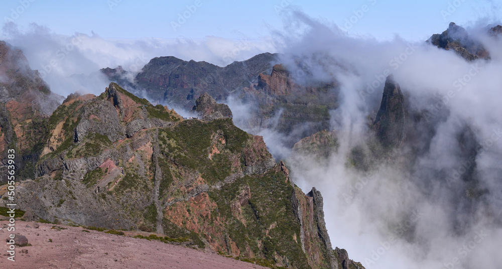 Panorama from Pico do Areeiro, a starting point of PR1 trail in Madeira Island. Hikers on trail to Pico Ruivo, the highest in Madeira. Fog ascending from a valley and remaining among mountain folds.