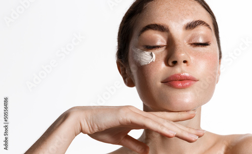 Young woman with eyes closed, applies facial daily cream, cosmetic clay mask or moisturizing scrub on her healthy, natural face, stands over white background