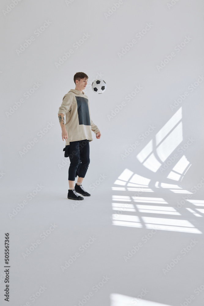 Young tattooed man in street style clothes kicking up soccer ball in spacious white hall illuminated with sunlight Young tattooed man in street style clothes kicking up soccer ball in spacious white hall illuminated with sunlight