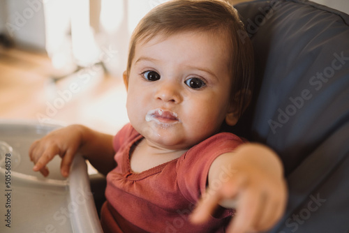 Baby girl sitting at the table for children with dirty mouth pointing with index finger to the camera.