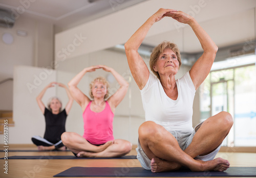 Sporty elderly woman meditating in yoga position Padmasana during group training at gym. Senior generation healthy lifestyle concept.