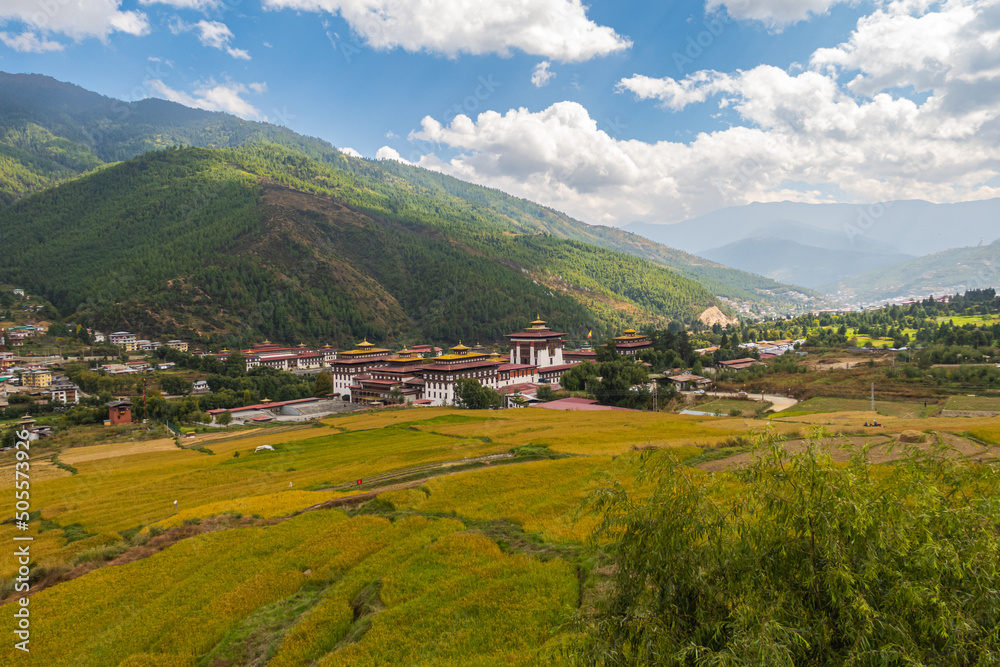 Wide angle panorama over the free wild open landscape of Bhutan. Punakha Dzong on the horizon. Meadows and agricultural land alternate. Clouds climb over the mountain. Valley with the Punakha Dzong