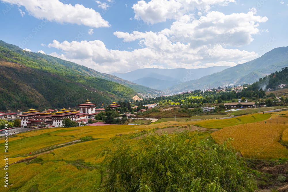 Wide angle panorama over the free wild open landscape of Bhutan. Punakha Dzong on the horizon. Meadows and agricultural land alternate. Clouds climb over the mountain. Valley with the Punakha Dzong