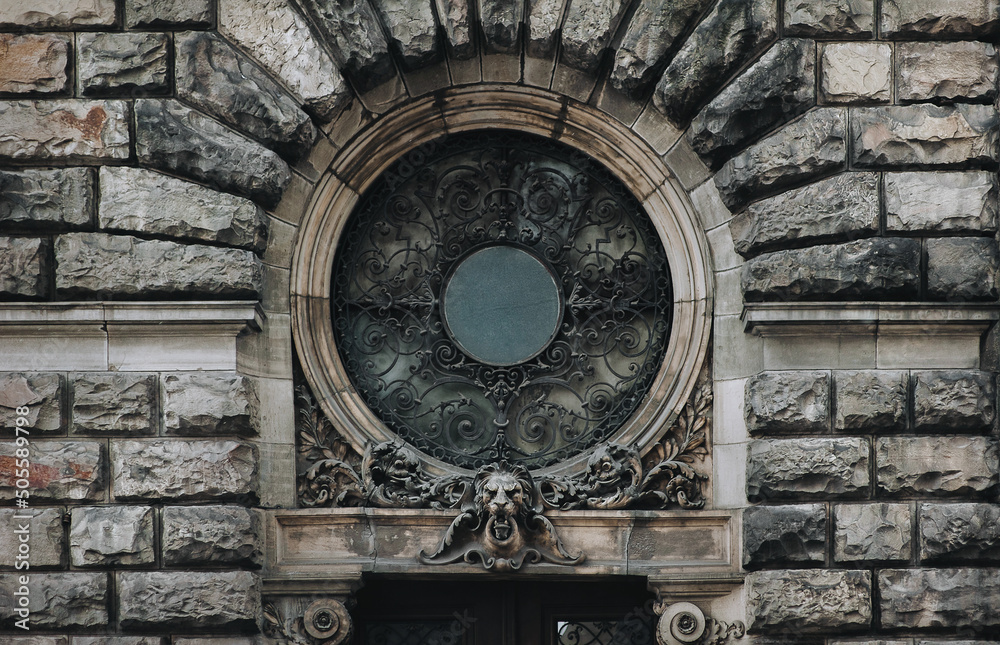 A round window with an openwork forged metal grill on the facade of an old house with stucco and decorative relief elements and a lion's head above the doorway. Lviv, Ukraine.