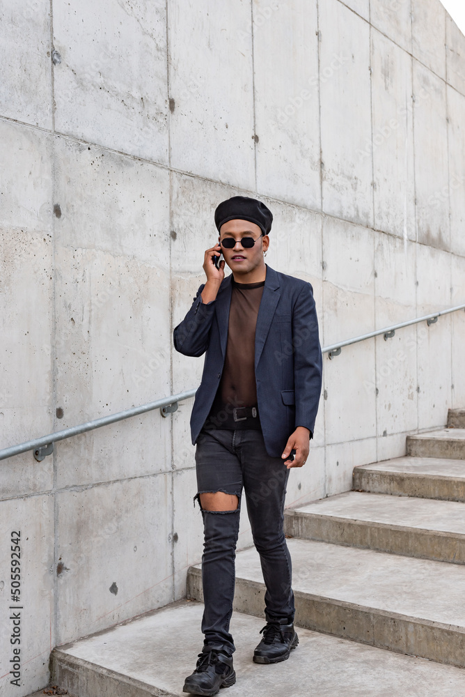Young Latin gay man wearing a fashionable hat and sunglasses, walking down the stairway while talking on his smartphone, isolated on a white background. LGTB. Copy space