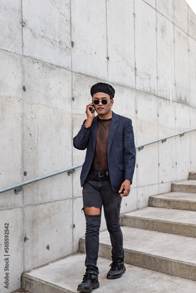 Young Latin gay man wearing a fashionable hat and sunglasses, walking down the stairway while talking on his smartphone, isolated on a white background. LGTB. Copy space Young Latin gay man wearing a fashionable hat and sunglasses, walking down the stairway while talking on his smartphone, isolated on a white background. LGTB. Copy space