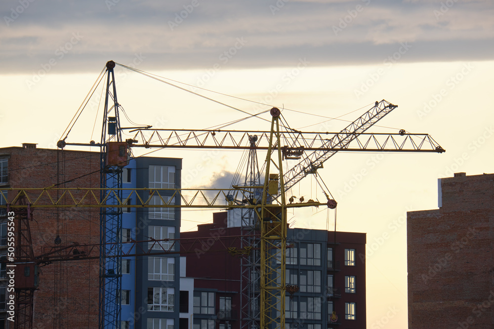 Dark silhouette of tower cranes at high residential apartment buildings construction site at sunset. Real estate development Dark silhouette of tower cranes at high residential apartment buildings construction site at sunset. Real estate development