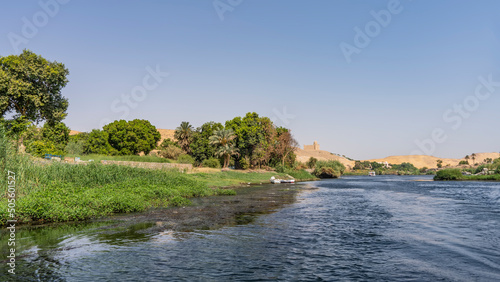 The bed of the Nile River bends. Green vegetation grows on the banks. At the top of a sand dune  against a clear sky  the Aga Khan mausoleum is visible. Ripples on the blue water. Egypt. Aswan