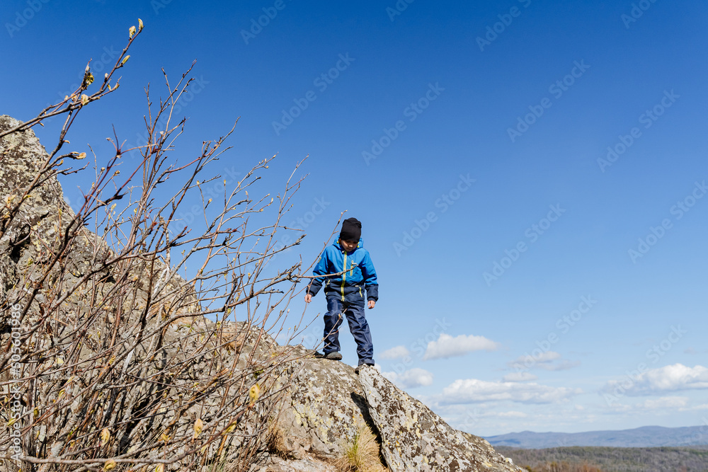 A child stands on a cliff high in the mountains, a trip during the summer holidays through the forest, a children's tourist camp, a young scout climbed a mountain, a child against the sky.,