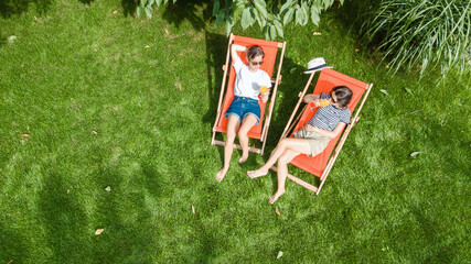 Young girls relax in summer garden in sunbed deckchairs on grass, women friends have fun outdoors in green park on weekend, aerial top view from above