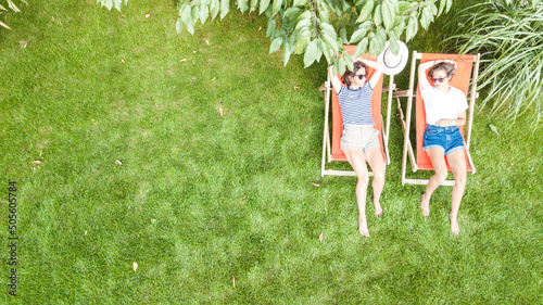 Fotografia Young girls relax in summer garden in sunbed deckchairs on grass, women friends