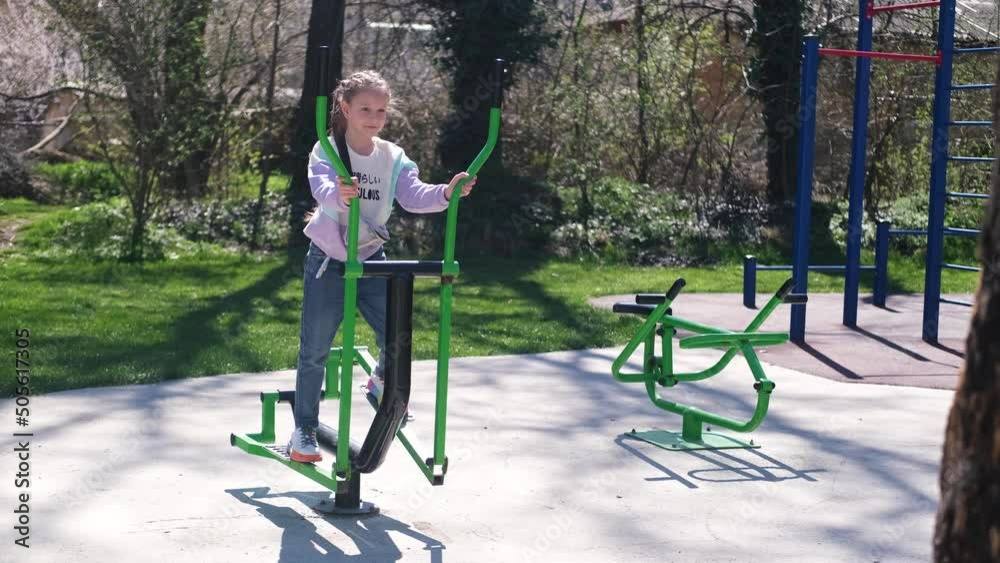 A little girl is engaged in a street sports ground. The child trains his legs on a city exercise bike. A little girl is engaged in a street sports ground. The child trains his legs on a city exercise bike.
