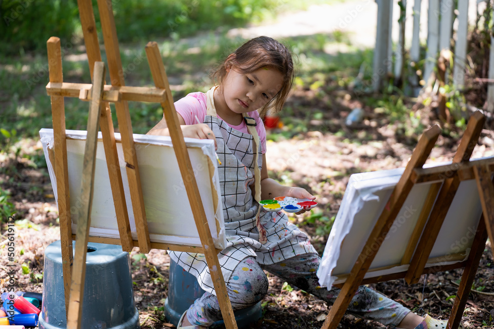 Φωτογραφία Little girl artist drawing a picture on canvas on an easel in nature, Cute little girl painting picture with a brush and color in backyard