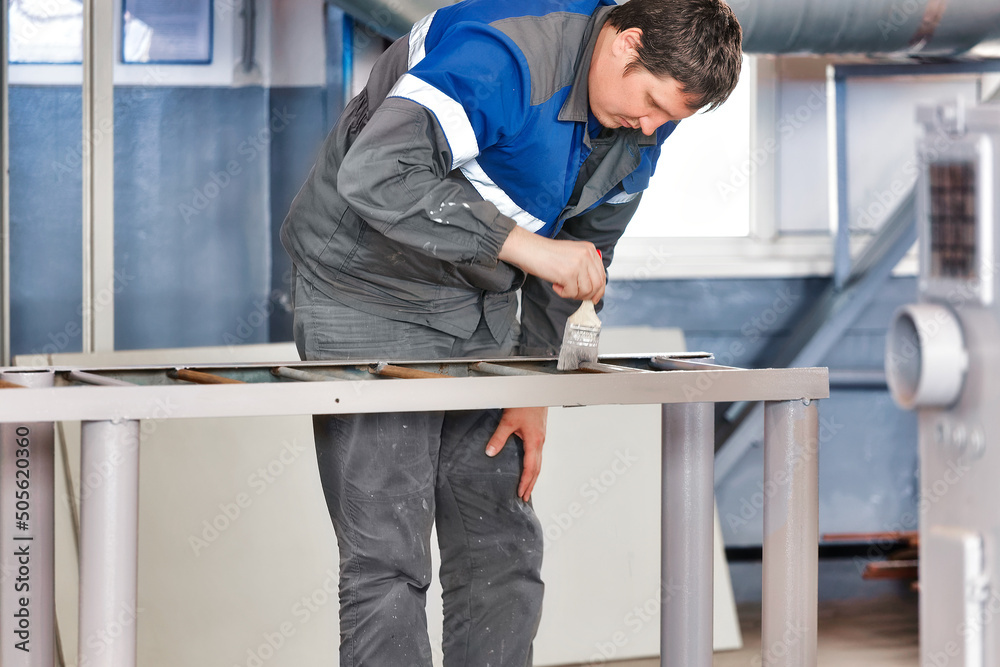 Caucasian worker in overalls paints metal rack or ladder with paint brush. Real workflow in production hall. Industrial painter at work.