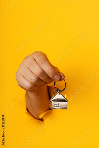 Male hand with metal house and key through a hole in yellow paper wall. House sale and rent concept. Vertical image