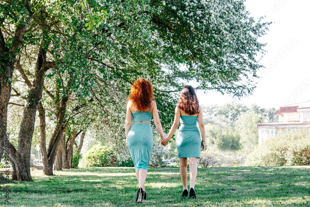A woman with red lush hair of European appearance holds the hand of a teenage girl with her back in the park under the trees. rear view of a mother and daughter walking on the street