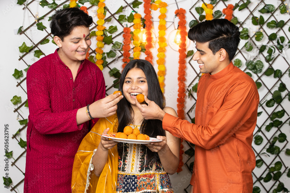 Portrait of happy indian young kids or bothers and sister wearing traditional cloths having fun eating laddu or laddoo sweets celebrating festival like diwali or rakshabandhan,
