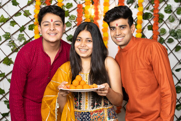 Portrait of happy indian young kids or bothers and sister wearing traditional cloths holding plate full of laddu or laddoo sweets celebrating festival like diwali looking at camera with smile.
