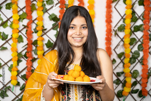 Portrait of happy indian teenage girl wearing traditional cloths holding plate full of laddu or laddoo sweets celebrating festival like diwali. photo