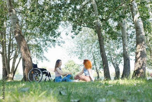 A woman with a disability and her female friend having a conversation in a shade of beautiful poplar trees, during a pleasant summer day