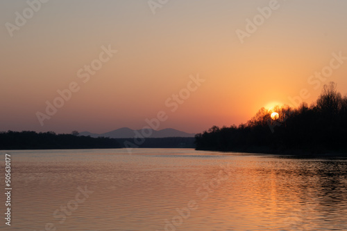 Landscape of river and mountantain before sunset, rippling water and distant mountain in haze with sun setting behind forest on clear sky, calming fair weather in nature