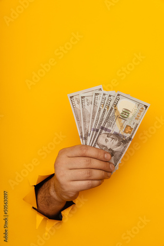 Hand holding money through hole in yellow paper wall. Vertical image