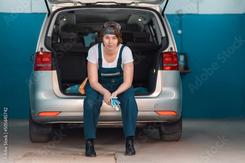 A female mechanic in coveralls is sitting in the open trunk of a car. Work at garage © _KUBE_