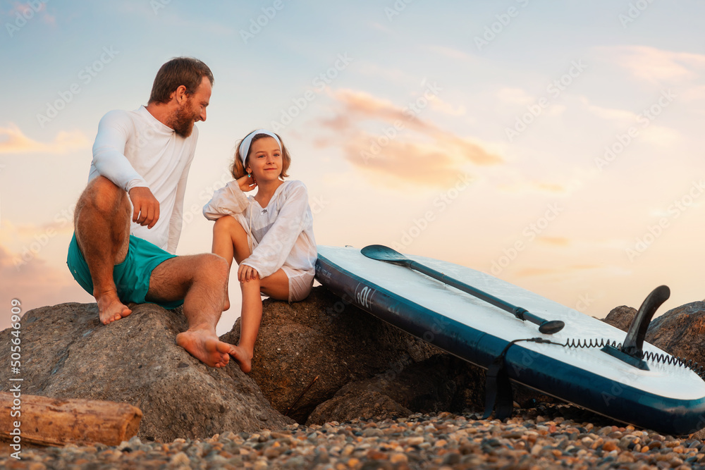 Summer activity vacations. Father and daughter sitting at the beach near sup board. Sunset sky at the background. Copy space