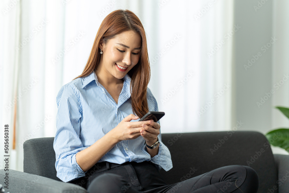 Smiling young asian woman using mobile phone while sitting on a couch at home, Technology and communication concept. Smiling young asian woman using mobile phone while sitting on a couch at home, Technology and communication concept.