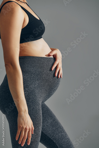 Cute pregnant belly isolated on gray. Side view of young pregnant woman embracing her abdomen with hands. Big belly on the third trimester of pregnancy close-up. Concept of pregnant life.