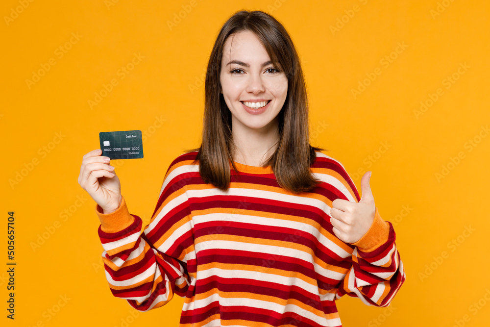 Φωτογραφία Young happy satisfied smiling caucasian woman 20s in red striped sweatshirt hold in hand credit bank card show thumb up isolated on plain yellow background studio portrait
