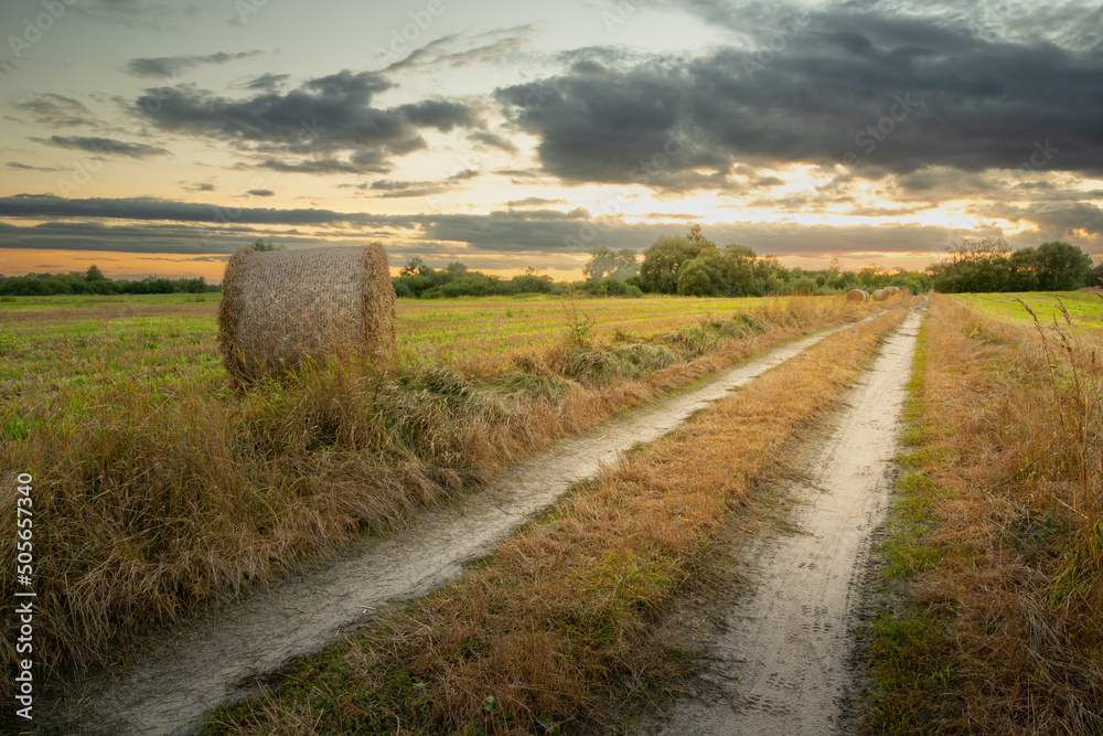 Country road and hay bale in the field, evening view with clouds