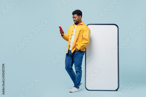 Full body man of African American ethnicity wearing yellow shirt stand big blank screen mobile cell phone with workspace copy space mockup area hold smartphone isolated on plain light blue background. photo