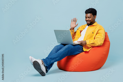 Full body young man of African American ethnicity wear yellow shirt sit in bag chair hold use work on laptop pc computer talk by video call waving hand isolated on plain pastel light blue background.