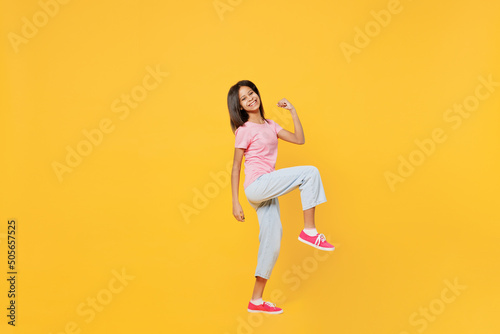 Full body little kid girl of African American ethnicity 12-13 years old in pink t-shirt doing winner gesture celebrate raise up leg isolated on plain yellow background. Childhood lifestyle concept