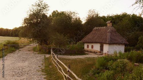 Small cute house surrounded by a simple wooden fence in rural area. Two little windows in house. The roof is thatched. Dense forest near the house.
