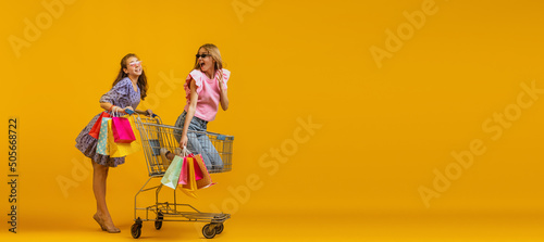 Portrait of cool young excited girls go shopping with shopping cart isolated on bright yellow background. Concept of sales, black friday, discount, emotions