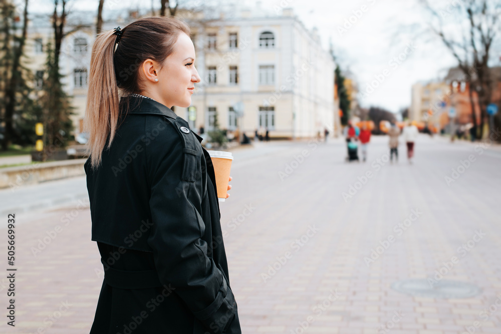Fototapeta premium Fashionable young woman in stylish coat holding coffee while walking in park and looking away. Back view of elegant caucasian blonde woman outdoors, copy space