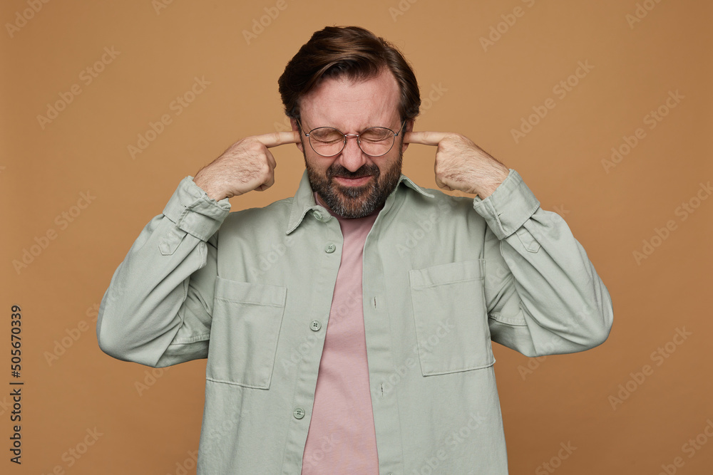 Studio portrait of bearded man standing over beige background wears casual shirt and glasses grimaced, plug ears with a fingers with irritated facial expression