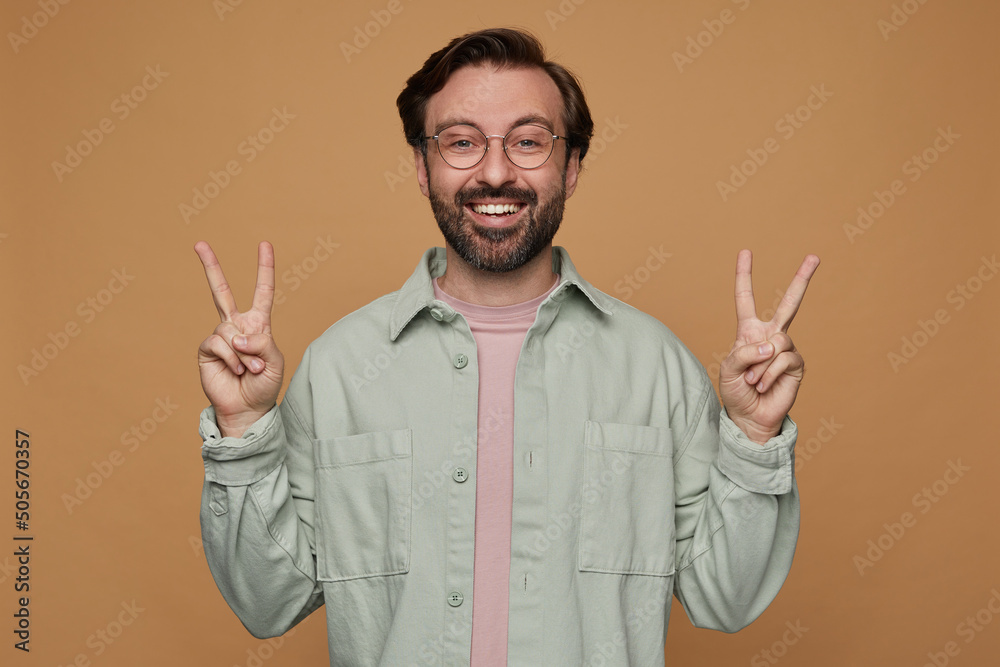 Studio portrait of bearded man standing over beige background wears casual shirt and glasses showing victory gesture into camera Studio portrait of bearded man standing over beige background wears casual shirt and glasses showing victory gesture into camera
