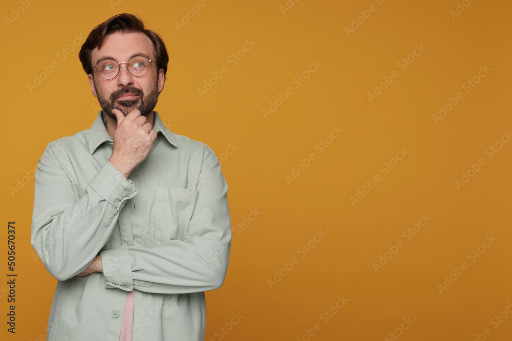Studio portrait of bearded man standing over orange background wears casual shirt and glasses thoughtfully looks aside, touching his chin