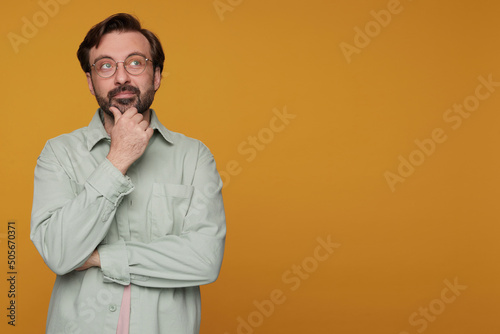 Studio portrait of bearded man standing over orange background wears casual shirt and glasses thoughtfully looks aside, touching his chin © timtimphoto