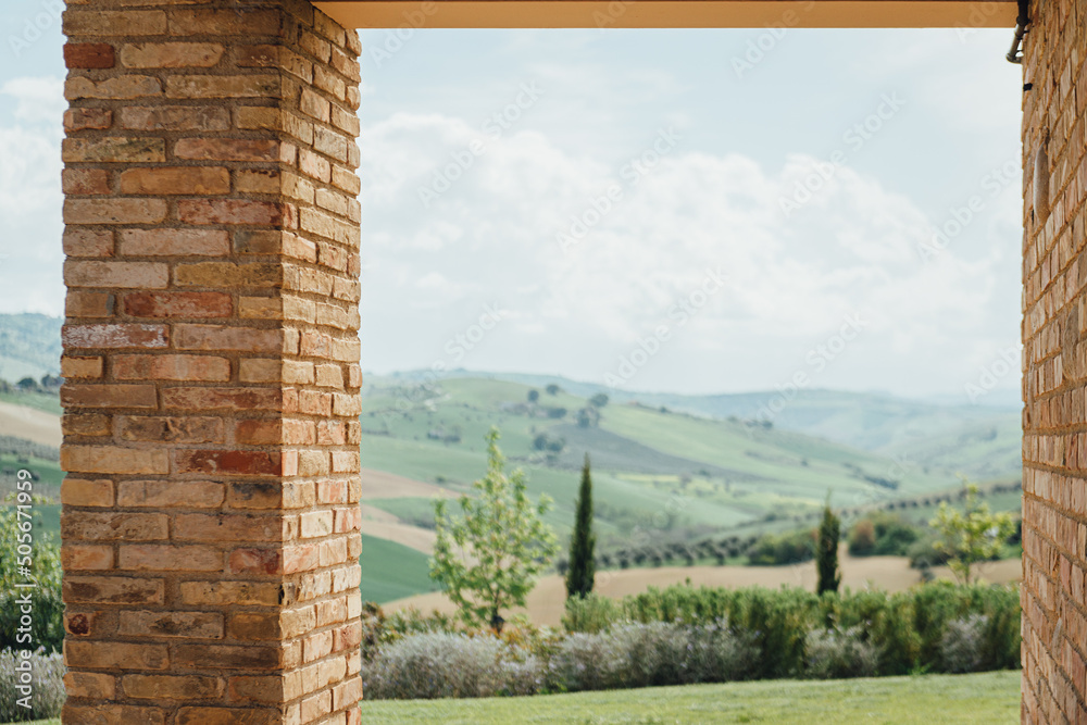 Fototapeta premium Italian traditional countryside view with hills and cypress trees, framed by brick columns and walls of modern villa