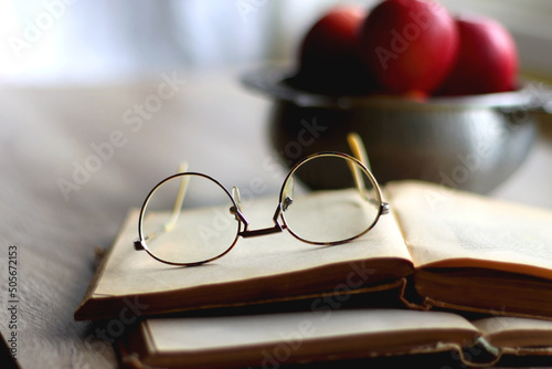 Silver bowl filled with red apples, open book and reading glasses on the table. Selective focus.