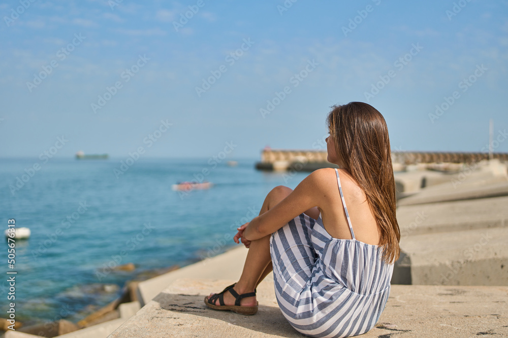 young European woman in a blue striped dress sitting on the harbour jetty enjoying the summer sun