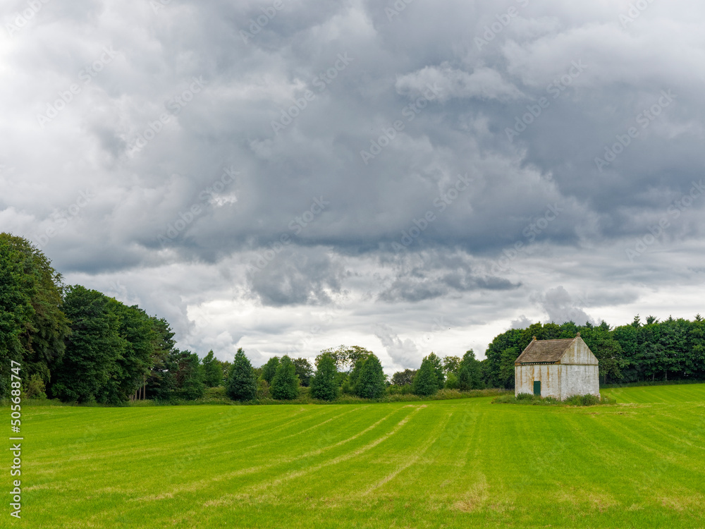 An Old well preserved white washed Doocot or Dovecote situated in a field of newly sown wheat with a mixed woodland behind and dark clouds above.