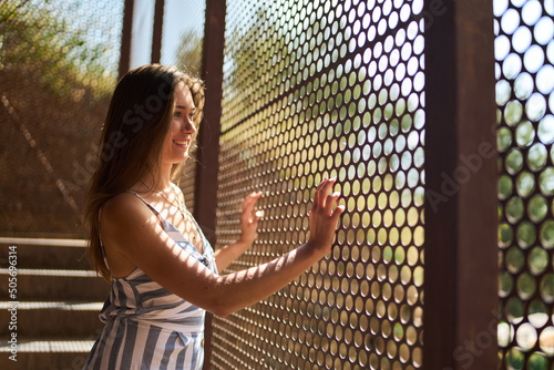 young woman posing next to a metallic mesh that lets the light shine through, bathing her skin with a pattern